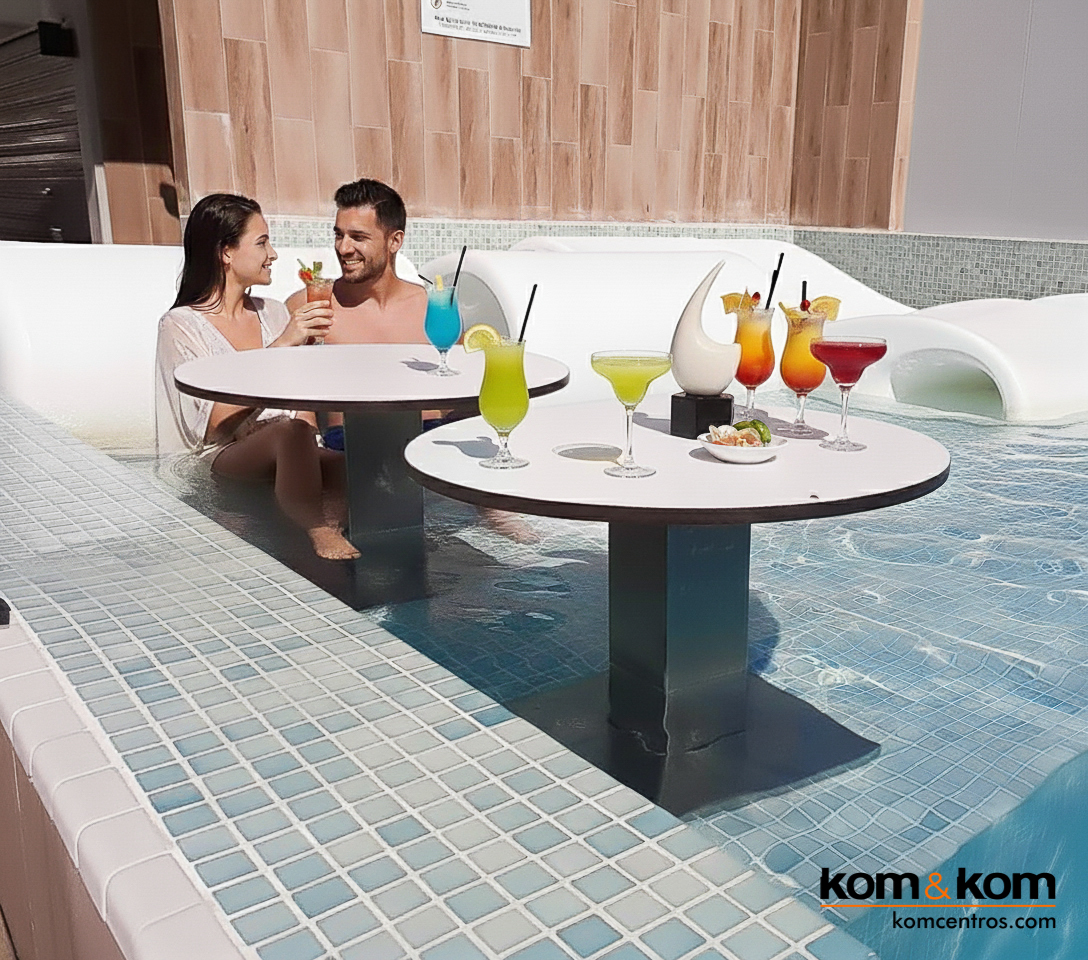 Pareja relaj&aacute;ndose en mesa dentro de piscina de hotel con c&oacute;cteles al atardecer. Terraza chill out de lujo con sombrillas blancas y ambiente elegante, estilo revista de decoraci&oacute;n.